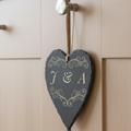 A heart-shaped dark slate hanging ornament with decorative scroll work and the initials 'J & A', suspended by a twine loop from a door knob on a light wood cabinet.