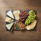A slate platter styled with cheeses, crackers, green and red grapes, nuts, and a small knife on a dark slate platter, set on a rustic wooden table.
