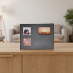 A rectangular black slate photo frame with three cutout slots, displayed on a light wood table in a softly blurred living room setting.