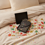 A slate gift box with a black lid and a small natural jute hanging loop, placed on top of a white gift bag printed with red and gold stars and swirls, on a rustic wooden surface with scattered confetti.