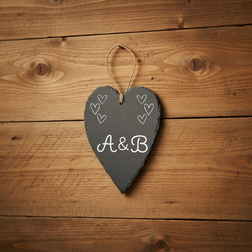 A heart-shaped dark gray slate hanging plaque with lettering reading 'A&B' in the center, small heart motifs near the top corners, and a natural twine loop at the top for hanging.